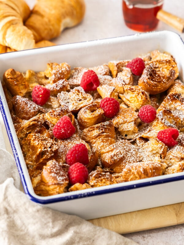 a white casserole dish with croissant bread pudding topped with powdered sugar and raspberries.