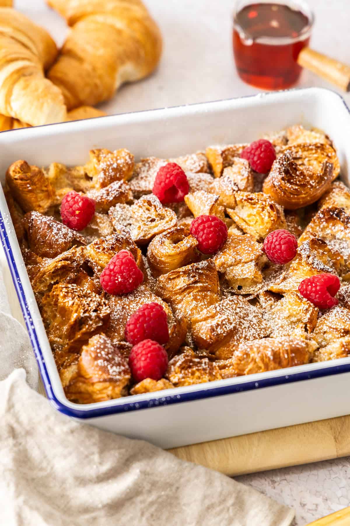 a white casserole dish with croissant bread pudding topped with powdered sugar and raspberries.