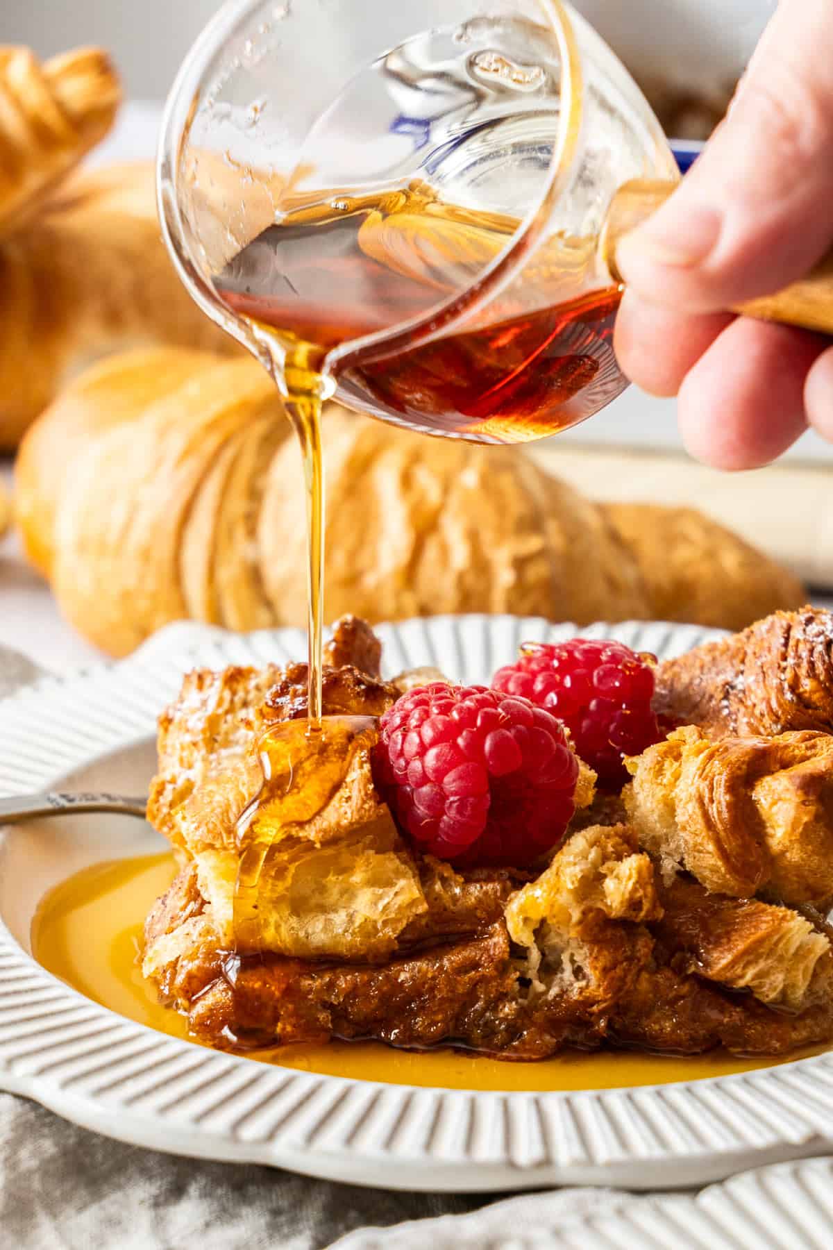 a hand pouring maple syrup over a plate of croissant casserole.