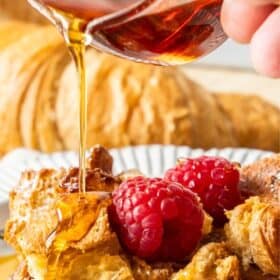 a hand pouring maple syrup over a plate of croissant casserole.