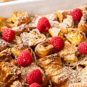 a white casserole dish with croissant bread pudding topped with powdered sugar and raspberries.