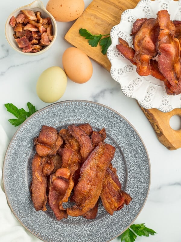 crispy air fryer bacon on a grey plate with a white plate with bacon, eggs, and parsley on a marbled board.