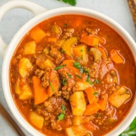 a white bowl of ground beef and vegetable soup topped with parsley, and a spoon, bread, and parsley on the side.