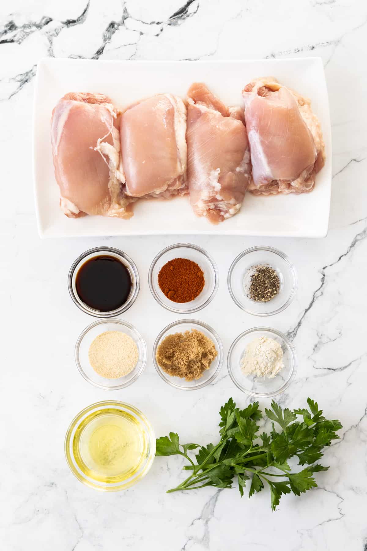 raw chicken thighs, parsley, olive oil, and spices on a marbled board.