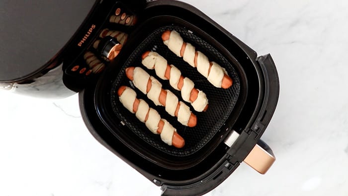 4 photos showing unbaked pigs in a pretzel blanket in an air fryer basket.