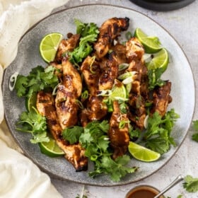 a grey plate of grilled peanut butter chicken topped with cilantro and lime wedges near bowls of peanut sauce and rice.