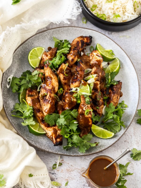 a grey plate of grilled peanut butter chicken topped with cilantro and lime wedges near bowls of peanut sauce and rice.