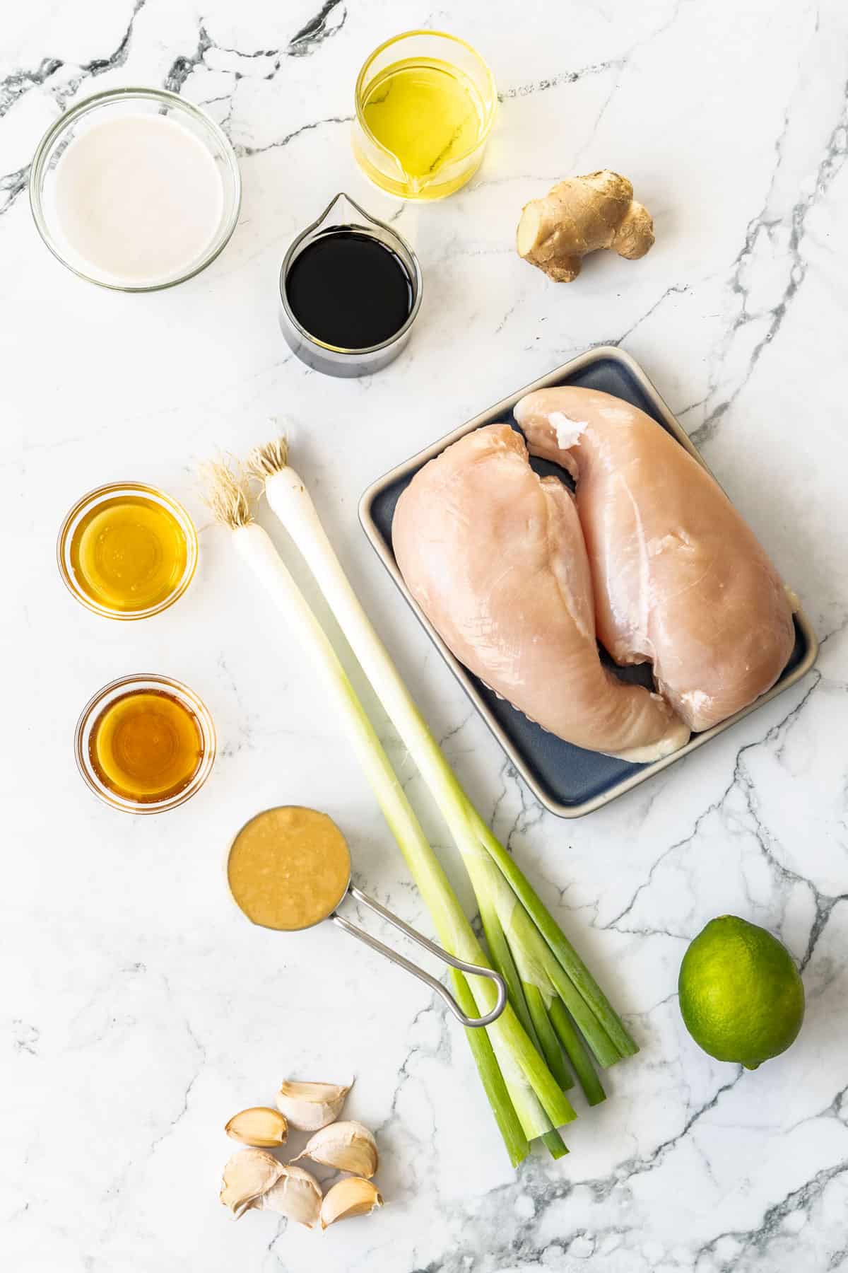 chicken breasts, ginger, lime, green onions, peanut butter, and other items on a marbled board.