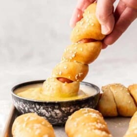 a hand dipping a pretzel dog in a dish of mustard.