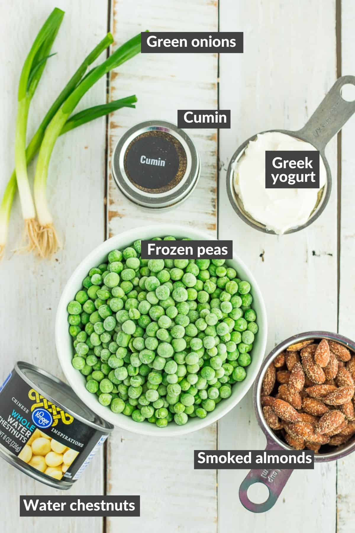 A bowl of peas, smoked almonds, green onions, Greek yogurt, cumin, and water chestnuts on a white wooden board.