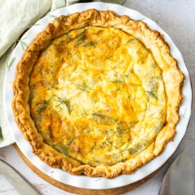 A delicious puff pastry quiche in a pie plate topped with fresh dill, with a napkin, utensils, and more dill on a marbled board.