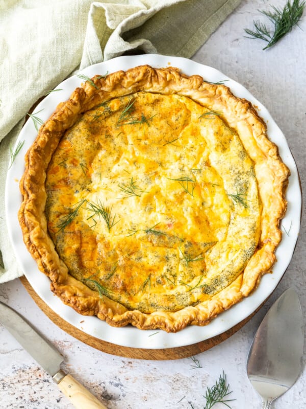 A delicious puff pastry quiche in a pie plate topped with fresh dill, with a napkin, utensils, and more dill on a marbled board.