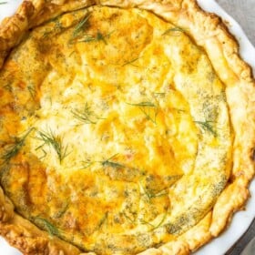 A delicious puff pastry quiche in a pie plate topped with fresh dill, with a napkin, utensils, and more dill on a marbled board.