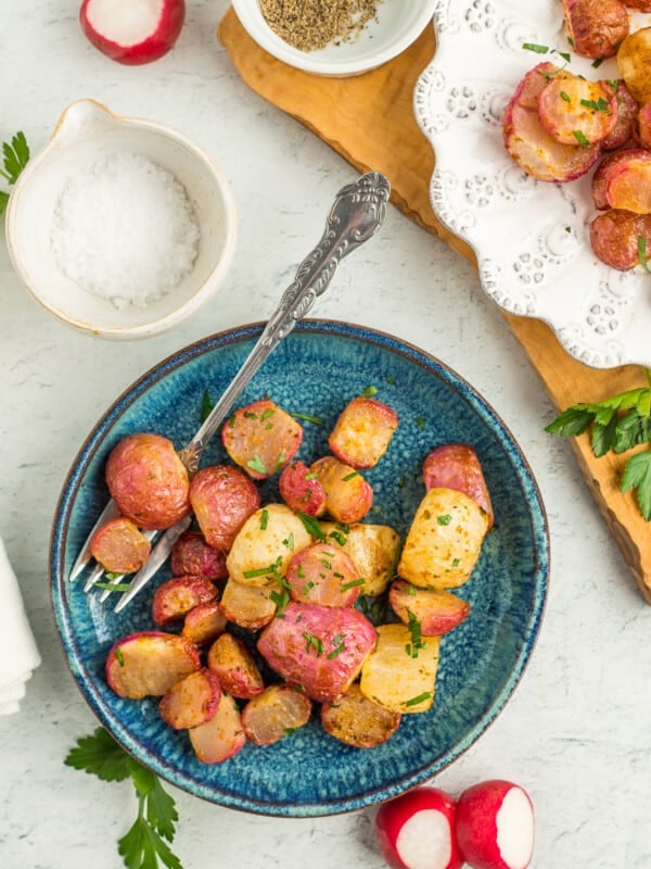 a teal plate with air fryer roasted radishes, fresh radishes, and a small dish of salt.