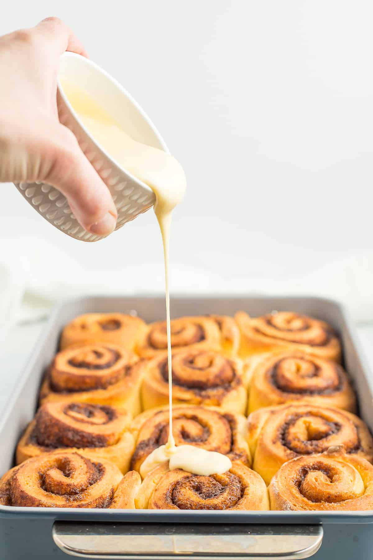 a hand pouring icing over a pan of cinnamon rolls.