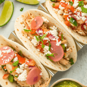 3 soft tacos on a grey board with lots of toppings, limes, etc.