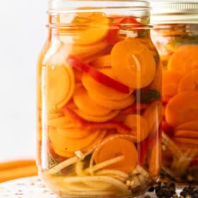 two jars of quick pickled carrots on a white board with peppercorns.