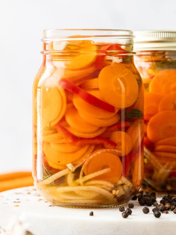 two jars of quick pickled carrots on a white board with peppercorns.