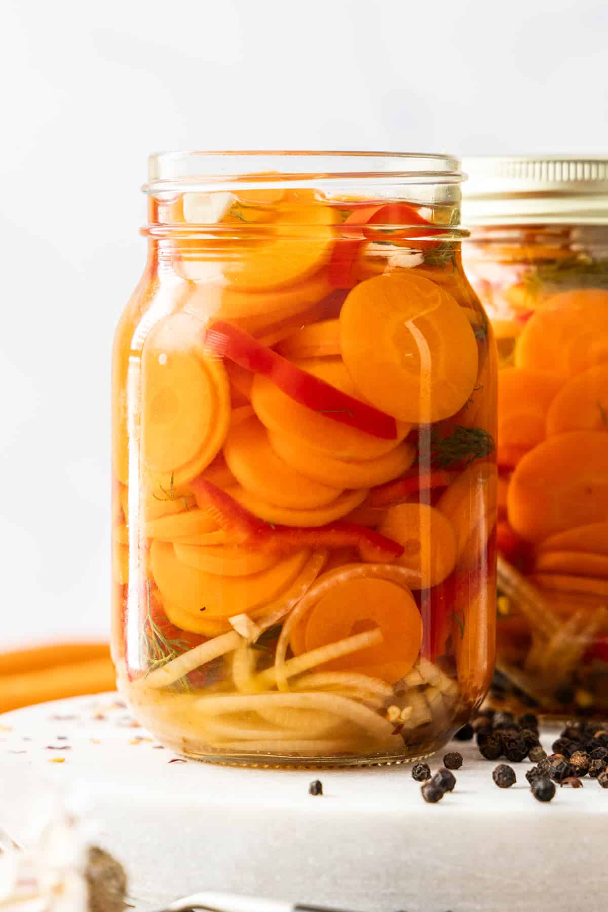 two jars of quick pickled carrots on a white board with peppercorns.