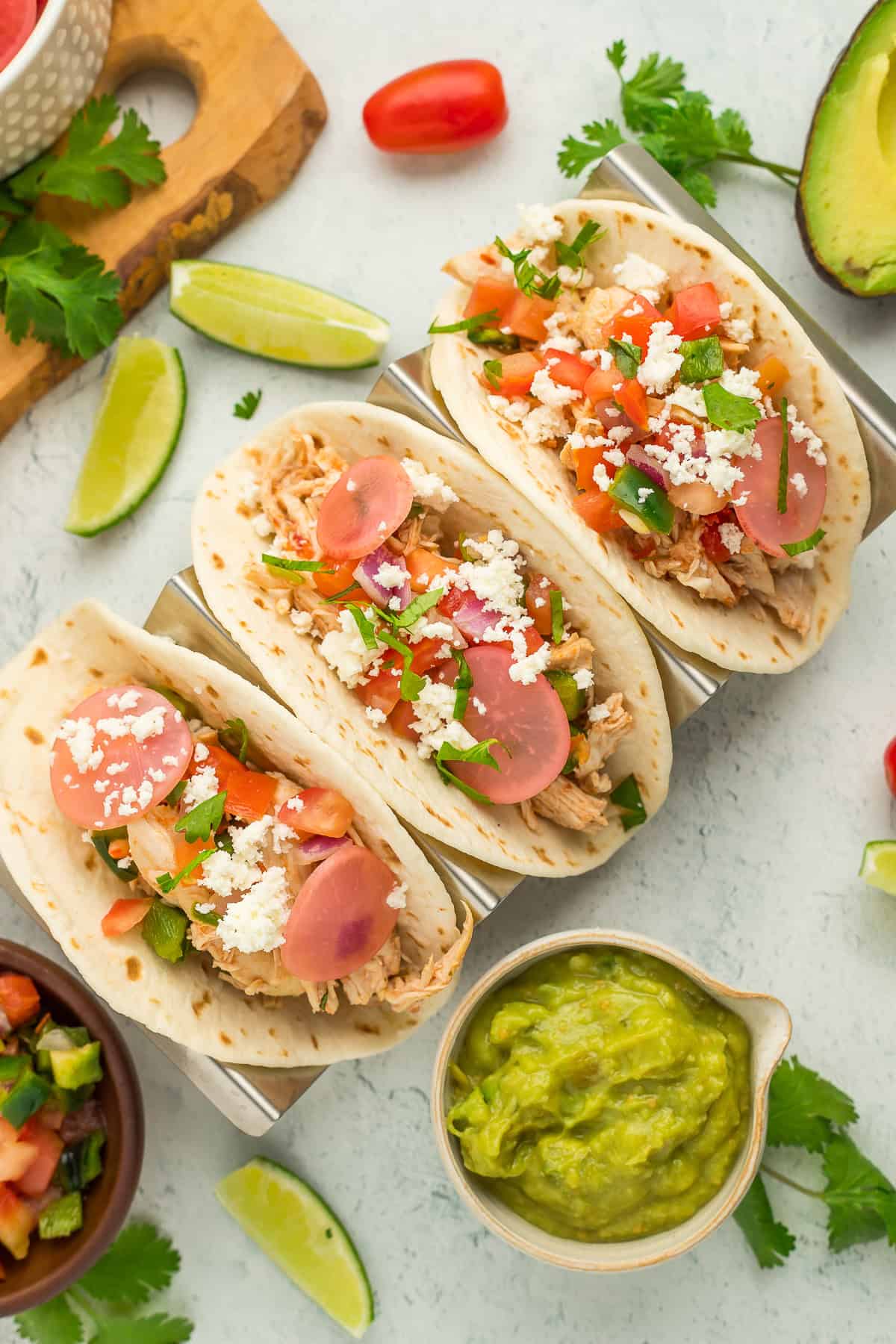 3 soft tacos on a grey board with lots of toppings, limes, etc.