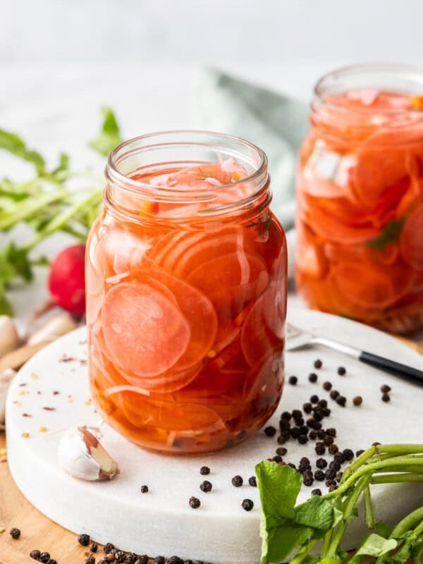 A canning jar of pickled radishes on a white board with peppercorns and radishes.