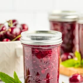 3 jars of cherry jam with fresh cherries, a bowl of cherries, and mint.