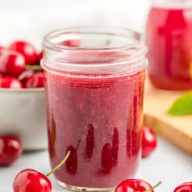 Two jars of cherry syrup on a white surface with a bowl of fresh cherries.