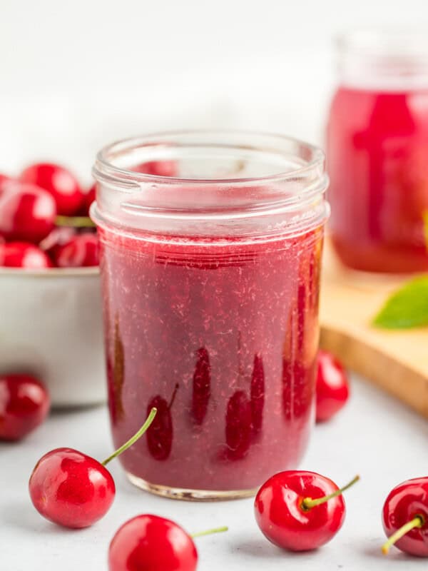 Two jars of cherry syrup on a white surface with a bowl of fresh cherries.