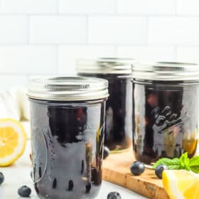 3 jars of canned blueberries against a tile wall with fresh blueberries, a lemon, and mint.