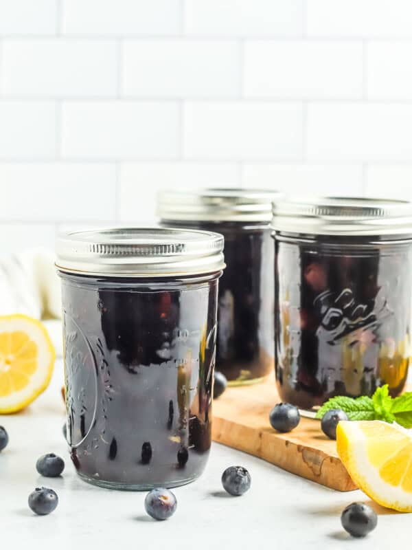3 jars of canned blueberries against a tile wall with fresh blueberries, a lemon, and mint.
