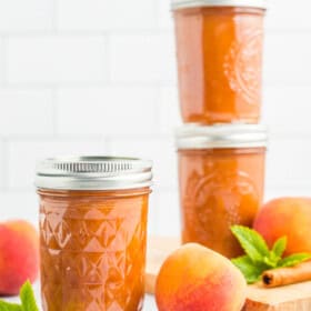 3 jars of peach butter with fresh peaches, cinnamon sticks, and mint against a tile background.