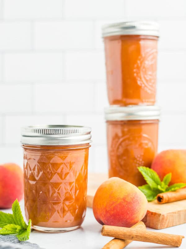 3 jars of peach butter with fresh peaches, cinnamon sticks, and mint against a tile background.