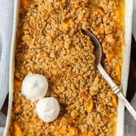 A white baking dish of peach crisp with a spoon and two scoops of ice cream.