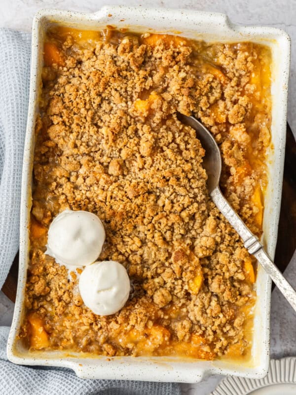 A white baking dish of peach crisp with a spoon and two scoops of ice cream.