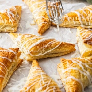 puff pastry apple turnovers with a whisk drizzling glaze over the top.