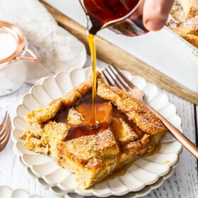 a hand pouring a glass container of maple syrup over a serving of brioche French toast casserole on a white plate.