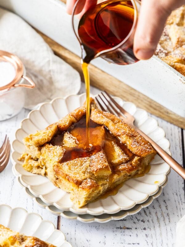 a hand pouring a glass container of maple syrup over a serving of brioche French toast casserole on a white plate.