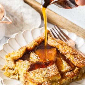 a hand pouring a glass container of maple syrup over a serving of brioche French toast casserole on a white plate.