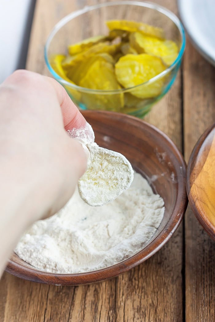 a hand dipping a pickle into a bowl of flour.
