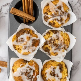 an overhead photo of 5 cinnamon roll muffins in a baking tray with cinnamon sticks on one of the parts of the pan.