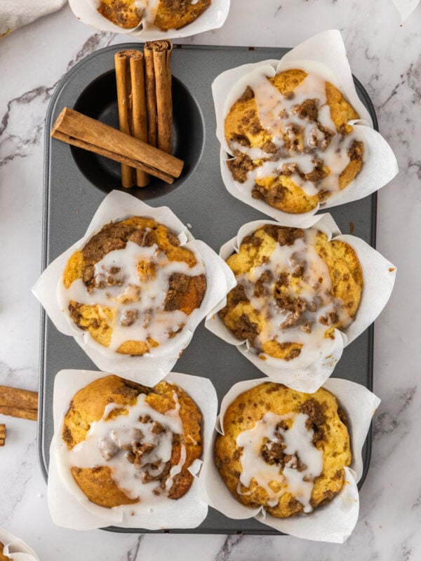 an overhead photo of 5 cinnamon roll muffins in a baking tray with cinnamon sticks on one of the parts of the pan.
