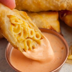 a hand dipping a mac and cheese egg roll into a bowl of buffalo ranch.