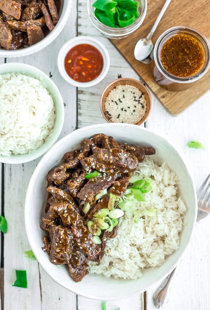 a white bowl of rice and beef teriyaki with bowls of sesame seeds, rice, hot sauce, and teriyaki sauce.