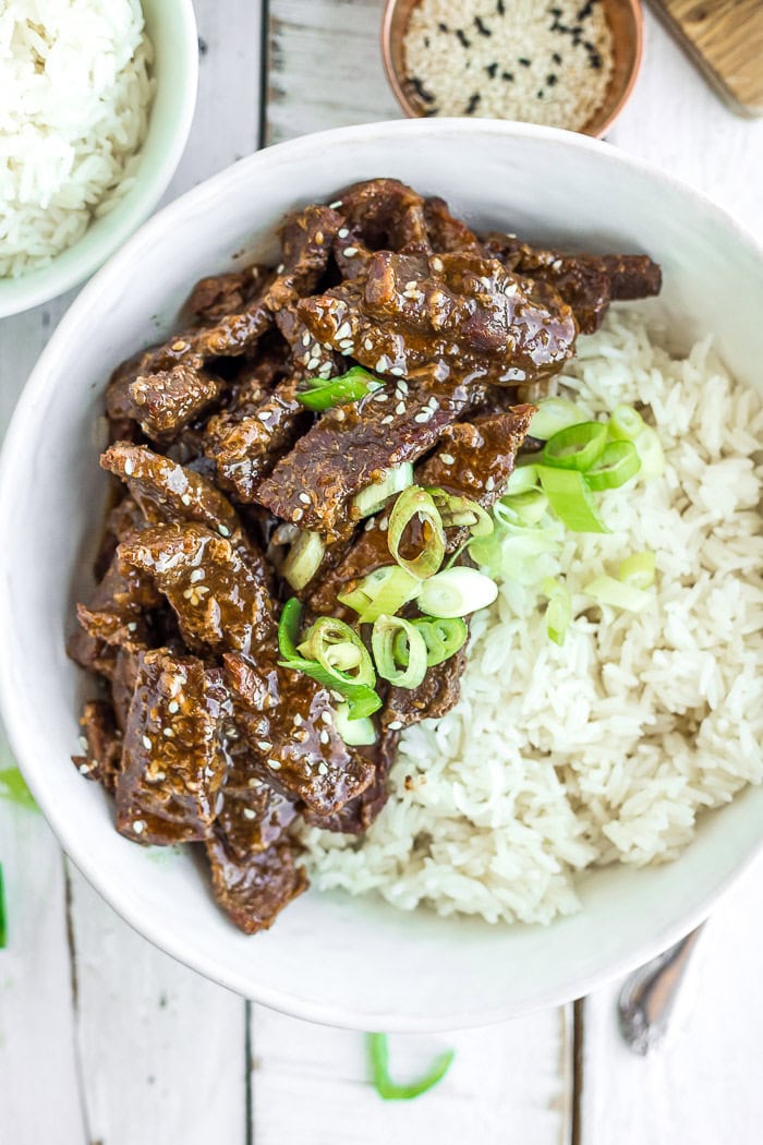 a white bowl of sliced Instant Pot teriyaki beef with rice topped with sesame seeds and green onions.