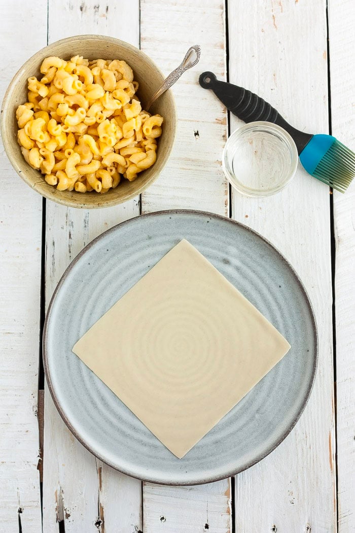 a bowl of macaroni and cheese, a bowl of water with a pastry brush, and an egg roll wrapper on a grey plate.