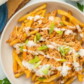 A white bowl with buffalo chicken fries with ranch drizzled over the top, and a small bowl of buffalo sauce.