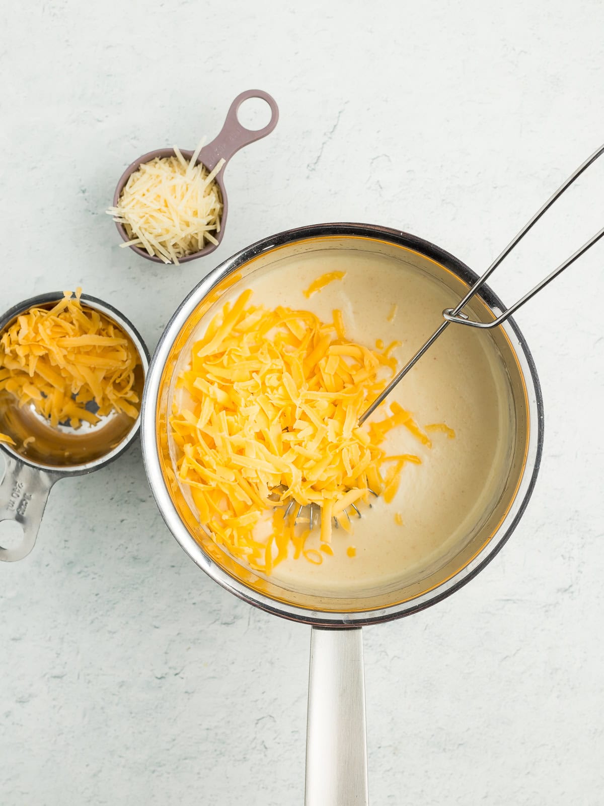 a whisk in a saucepan with a white sauce and shredded cheese.