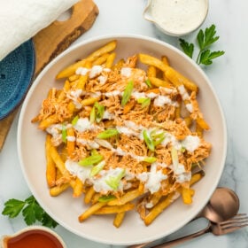 A white bowl with buffalo chicken fries with ranch drizzled over the top, and a small bowl of buffalo sauce.