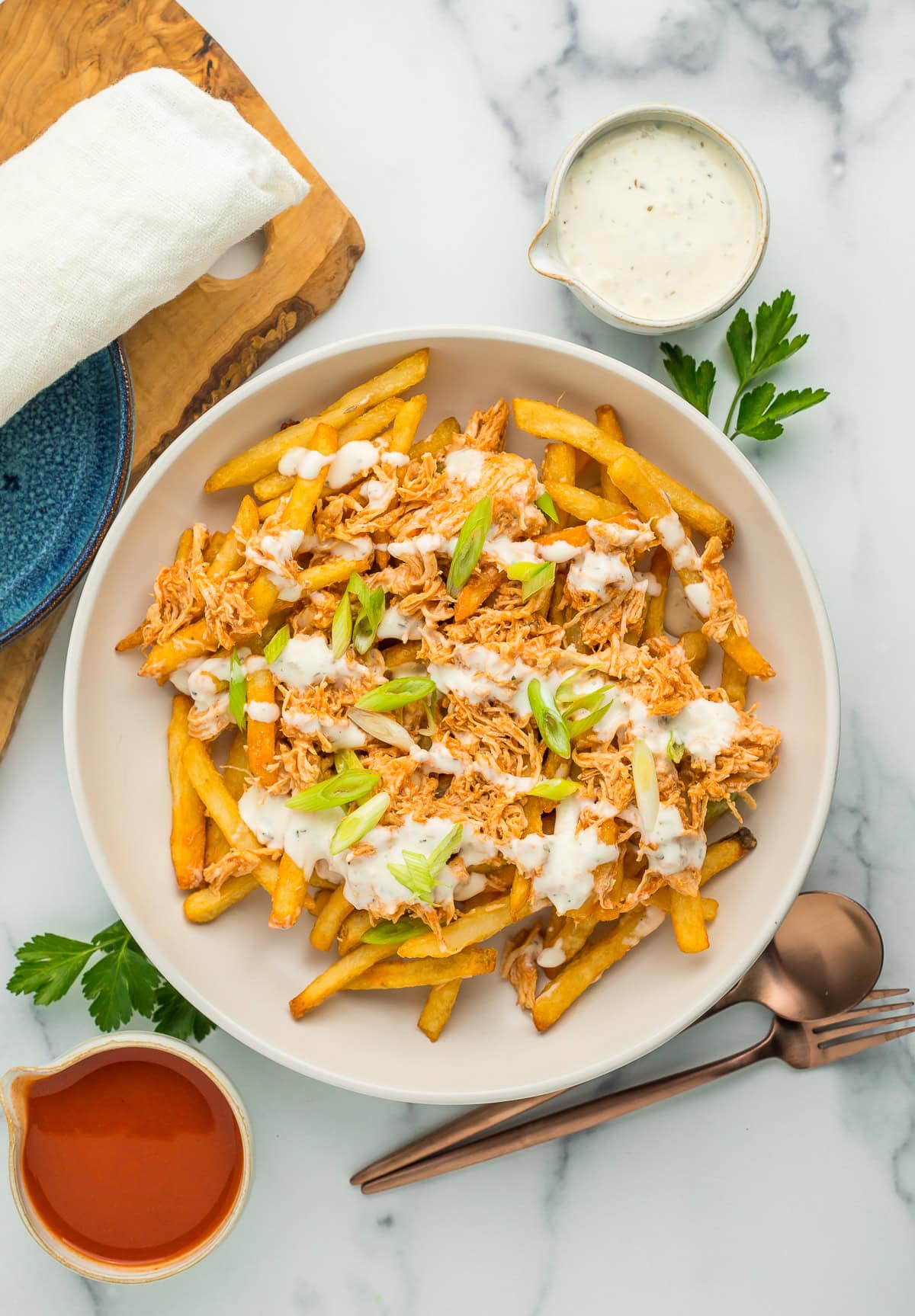 A white bowl with buffalo chicken fries with ranch drizzled over the top, and a small bowl of buffalo sauce.