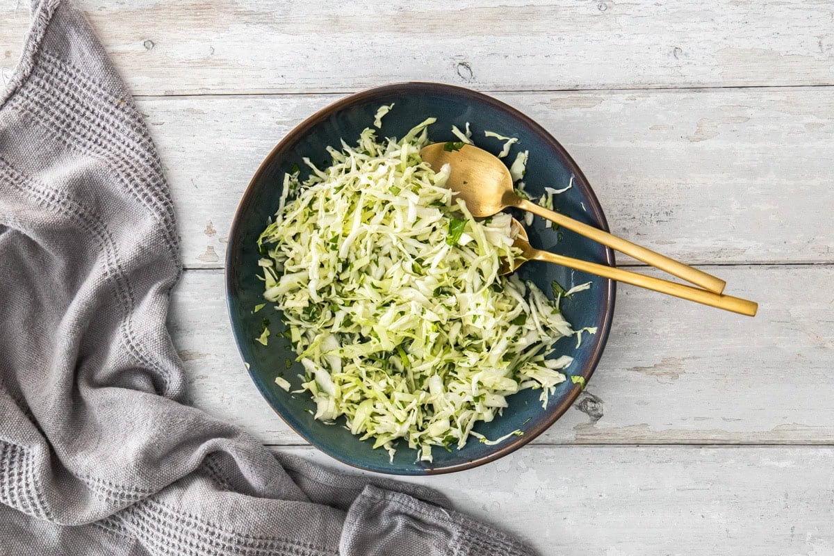A dark blue bowl filled with cabbage slaw and two spoons.
